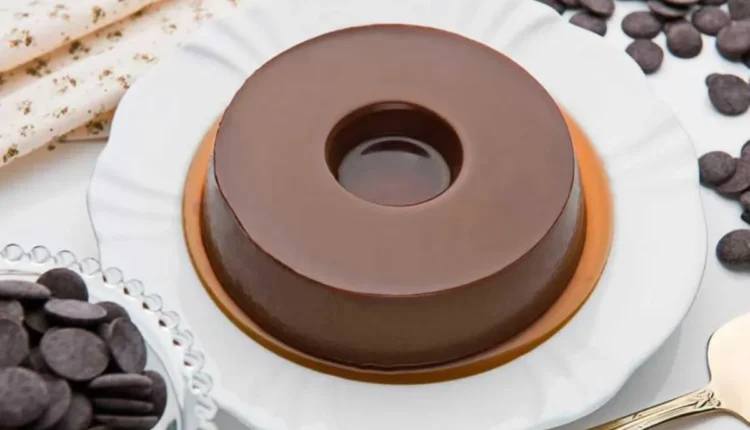 Pudim de chocolate com formato circular, servido em prato branco com borda dourada, ao lado de chocolate em pedaços.