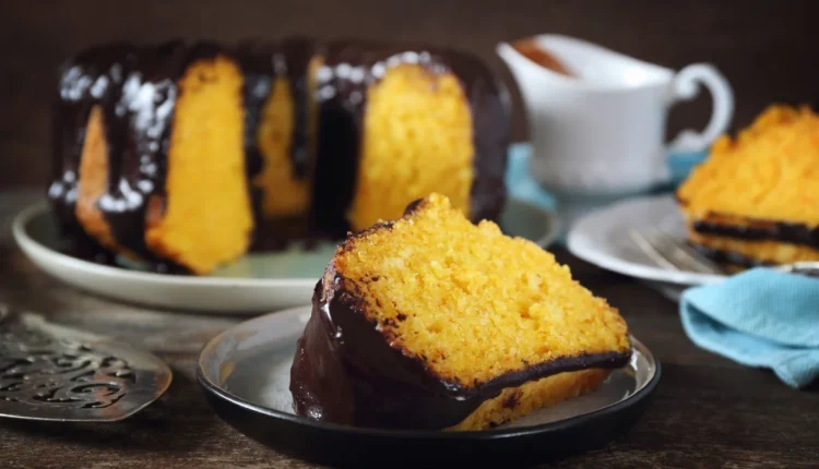 Fatia de bolo de cenoura macio com cobertura de chocolate, servida em prato, com bolo inteiro ao fundo