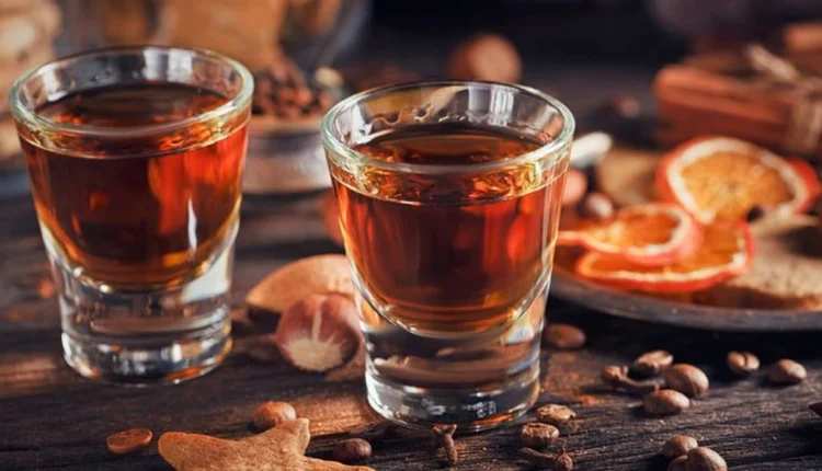 Dois copos pequenos com licor de jenipapo, cercados por especiarias e frutas secas em mesa de madeira.