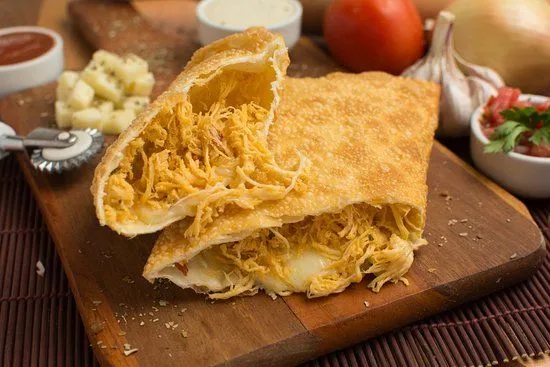 Pastel de frango desfiado com queijo Pastel crocante recheado com frango desfiado e queijo derretido, servido sobre uma tábua de madeira com ingredientes ao fundo.