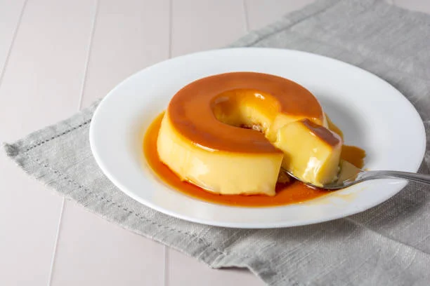 Pudim de Leite Cremoso e Irresistível Pudim de leite cremoso com calda dourada, servido em prato branco, com pedaço sendo retirado